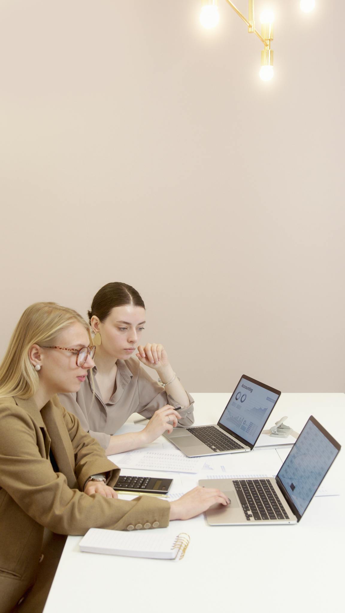 Two professional women collaborating on laptops in a modern office setting.